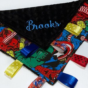 Op de afbeelding: Een zwart-witte babydeken met een superhelden-thema en een zwarte minky-rug. De deken heeft verschillende gekleurde satijnen linten die aan de randen zijn bevestigd. De deken is gepersonaliseerd met de naam "Brooks" geborduurd in blauwe draad.