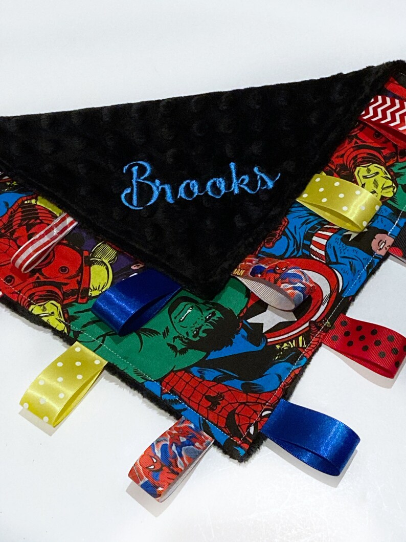 Op de afbeelding: Een zwart-witte babydeken met een superhelden-thema en een zwarte minky-rug. De deken heeft verschillende gekleurde satijnen linten die aan de randen zijn bevestigd. De deken is gepersonaliseerd met de naam "Brooks" geborduurd in blauwe draad.