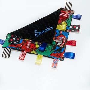 Op de afbeelding: Een zwarte en blauwe babydeken met een superhelden-thema en een zwarte minky-rug. De deken heeft verschillende gekleurde linten aan de randen en is gepersonaliseerd met de naam "Brooks" geborduurd in blauwe draad.