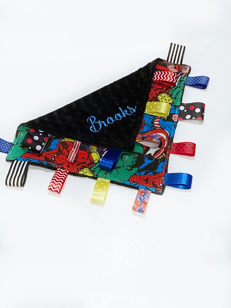 Op de afbeelding: Een zwarte en blauwe babydeken met een superhelden-thema en een zwarte minky-rug. De deken heeft verschillende gekleurde linten aan de randen en is gepersonaliseerd met de naam "Brooks" geborduurd in blauwe draad.