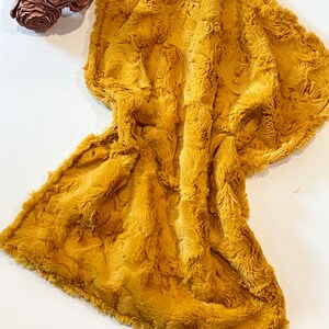 May include: A soft, plush mustard yellow throw blanket with a fuzzy texture. The blanket is folded and draped over a white surface.