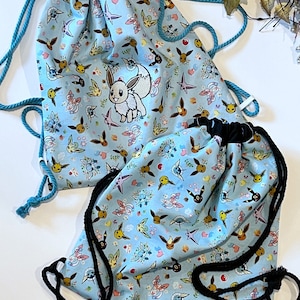 May include: Two blue drawstring bags with a white background and a repeating pattern of cartoon Pokemon characters. One bag has a larger embroidered image of a Pokemon character.