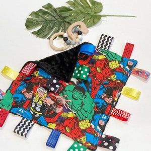 Może przedstawiać: Czarno-biały kocyk dla niemowląt z kolorowym nadrukiem superbohater&oacute;w. Kocyk ma wiele wszytych lam&oacute;wek w r&oacute;żnych kolorach i wzorach, w tym grochy, paski i zygzaki.