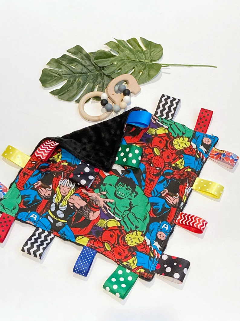 Może przedstawiać: Czarno-biały kocyk dla niemowląt z kolorowym nadrukiem superbohater&oacute;w. Kocyk ma wiele wszytych lam&oacute;wek w r&oacute;żnych kolorach i wzorach, w tym grochy, paski i zygzaki.