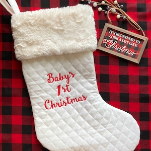 May include: White quilted Christmas stocking with a faux fur cuff. The stocking is embroidered with the text "Baby's 1st Christmas". A small wooden sign with the text "It's beginning to look a lot like Christmas" is in the background.