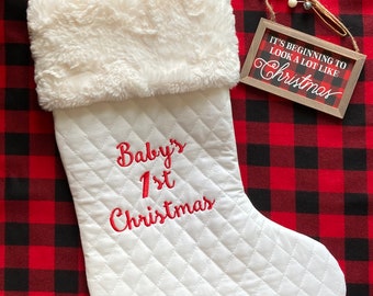 May include: White quilted Christmas stocking with a faux fur cuff. The stocking is embroidered with the text "Baby's 1st Christmas". A small wooden sign with the text "It's beginning to look a lot like Christmas" is in the background.