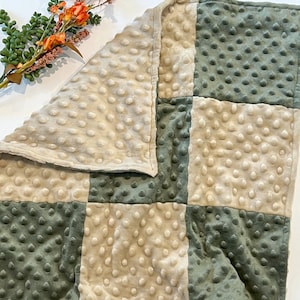 May include: A soft, quilted baby blanket with a patchwork design. The blanket is made of two colors: a light beige and a dark green. The blanket is made of a soft, plush fabric.
