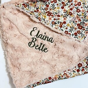 Może przedstawiać: Miękki, r&oacute;żowy kocyk dla dziecka z wyhaftowanym imieniem "Elaina Belle" zieloną nicią. Kocyk ma pluszową, teksturowaną powierzchnię i kwiatową podszewkę z czerwonymi, ż&oacute;łtymi i białymi kwiatami. Na wierzchu umieszczono ozdobny kwiat.