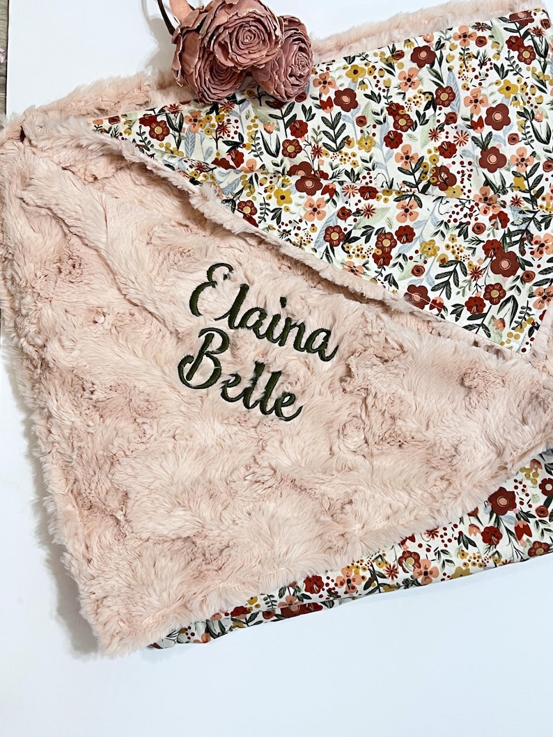 Może przedstawiać: Miękki, r&oacute;żowy kocyk dla dziecka z wyhaftowanym imieniem "Elaina Belle" zieloną nicią. Kocyk ma pluszową, teksturowaną powierzchnię i kwiatową podszewkę z czerwonymi, ż&oacute;łtymi i białymi kwiatami. Na wierzchu umieszczono ozdobny kwiat.