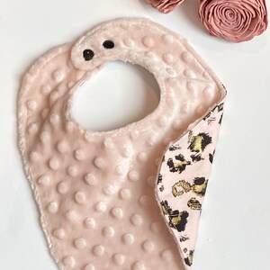 May include: A pink baby bib with a soft, plush texture on one side and a leopard print fabric on the other side. The bib has two black snaps for fastening.