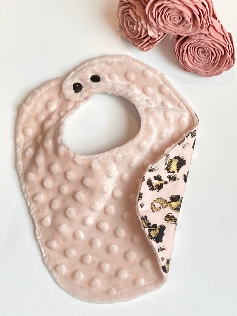 May include: A pink baby bib with a soft, plush texture on one side and a leopard print fabric on the other side. The bib has two black snaps for fastening.