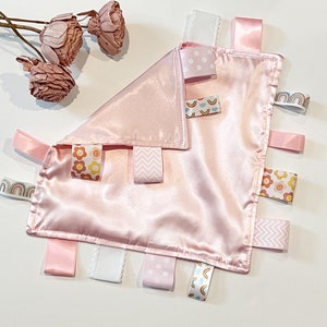 May include: A pink satin baby security blanket with various colorful ribbon tags. The tags feature patterns of rainbows, flowers, polka dots, and zigzags.