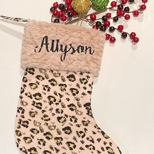 May include: A Christmas stocking with a pink leopard print pattern. The top of the stocking is a soft, light brown material with the name "Allyson" embroidered in black. Decorative red berries and gold and green glitter balls are placed above the stocking.