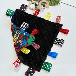 Może przedstawiać: Czarno-biały sensoryczny kocyk dla niemowląt z nadrukiem komiksu o superbohaterach po jednej stronie. Kocyk ma wiele kolorowych satynowych wstążek przymocowanych do krawędzi.