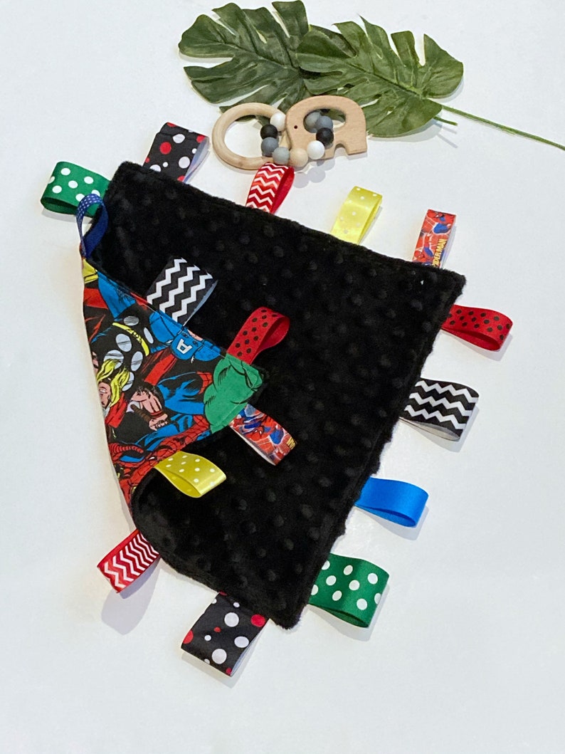 Może przedstawiać: Czarno-biały sensoryczny kocyk dla niemowląt z nadrukiem komiksu o superbohaterach po jednej stronie. Kocyk ma wiele kolorowych satynowych wstążek przymocowanych do krawędzi.