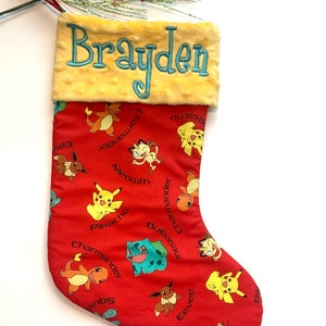 May include: A red Christmas stocking with a yellow cuff featuring the name "Brayden" embroidered in teal blue. The stocking is decorated with a repeating pattern of Pokemon characters, including Pikachu, Charmander, Bulbasaur, and Eevee.