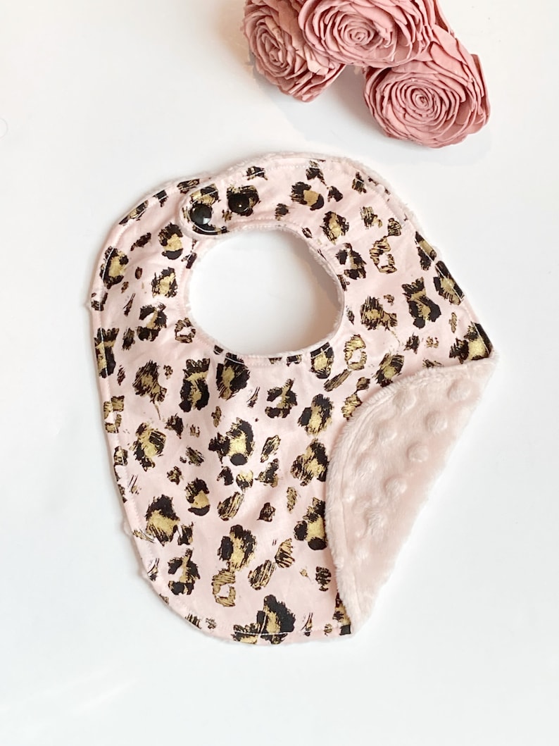 May include: Pink baby bib with a leopard print and gold accents. The bib has a soft, plush backing.