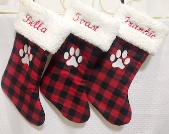 May include: Three red and black buffalo plaid Christmas stockings with white faux fur cuffs and a white paw print design. The stockings are personalized with the names "Bella", "Toast", and "Frankie".