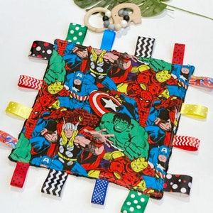 Może przedstawiać: Wielokolorowy kocyk bezpieczeństwa dla niemowląt z motywem superbohater&oacute;w. Kocyk przedstawia r&oacute;żnych superbohater&oacute;w, w tym Spidermana, Hulka, Kapitana Amerykę i Thora. Kocyk ma wiele wstążek przymocowanych do krawędzi w r&oacute;żnych kolorach i wzorach.