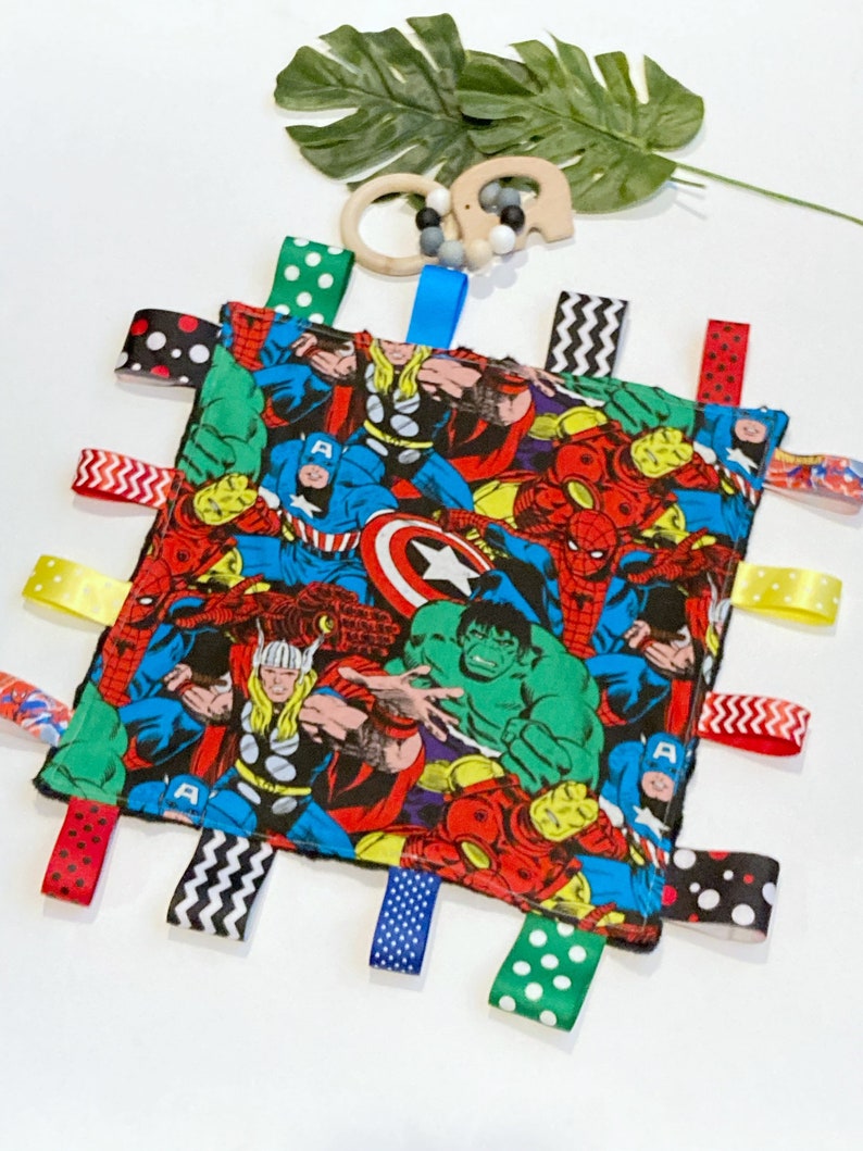 Może przedstawiać: Wielokolorowy kocyk bezpieczeństwa dla niemowląt z motywem superbohater&oacute;w. Kocyk przedstawia r&oacute;żnych superbohater&oacute;w, w tym Spidermana, Hulka, Kapitana Amerykę i Thora. Kocyk ma wiele wstążek przymocowanych do krawędzi w r&oacute;żnych kolorach i wzorach.