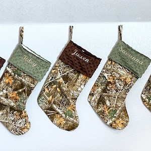 May include: Five personalized Christmas stockings with a camouflage pattern and brown or green fleece cuffs. The stockings are hung on a white wall and each has a name embroidered on the cuff: Weston, Jilly, Jaxen, Savanna, and Halen.