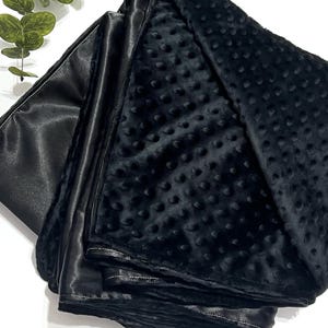 May include: A black blanket featuring a dual-sided design with one side made of smooth, solid black fabric and the other side composed of black minky fabric with a raised, textured dimpled pattern, finished with a stitched border edge.