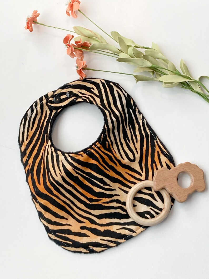 May include: A black and orange zebra print baby bib with two black snaps and a wooden teether ring with a flower-shaped wooden teether attached.