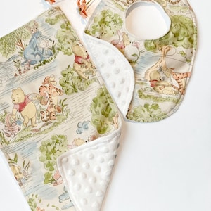 May include: A Winnie the Pooh-themed baby bib and burp cloth set. The bib is made of cotton fabric with a white minky backing. The burp cloth is made of the same cotton fabric with a white minky backing. Both feature a Winnie the Pooh and friends print.