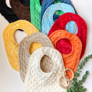 May include: Assortment of ten baby bibs in various colors, including brown, green, blue, red, yellow, orange, and beige. The bibs are made of soft, textured fabric with a dotted pattern.