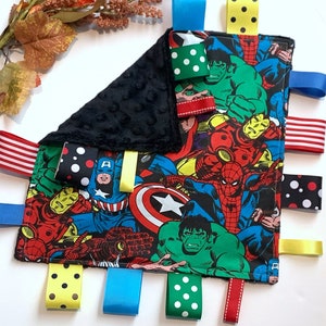 Op de afbeelding: Een zwart-wit babydeken met een kleurrijke superheldenprint. De deken heeft meerdere stoffen labels in verschillende kleuren en patronen, waaronder stippen, strepen en effen kleuren.