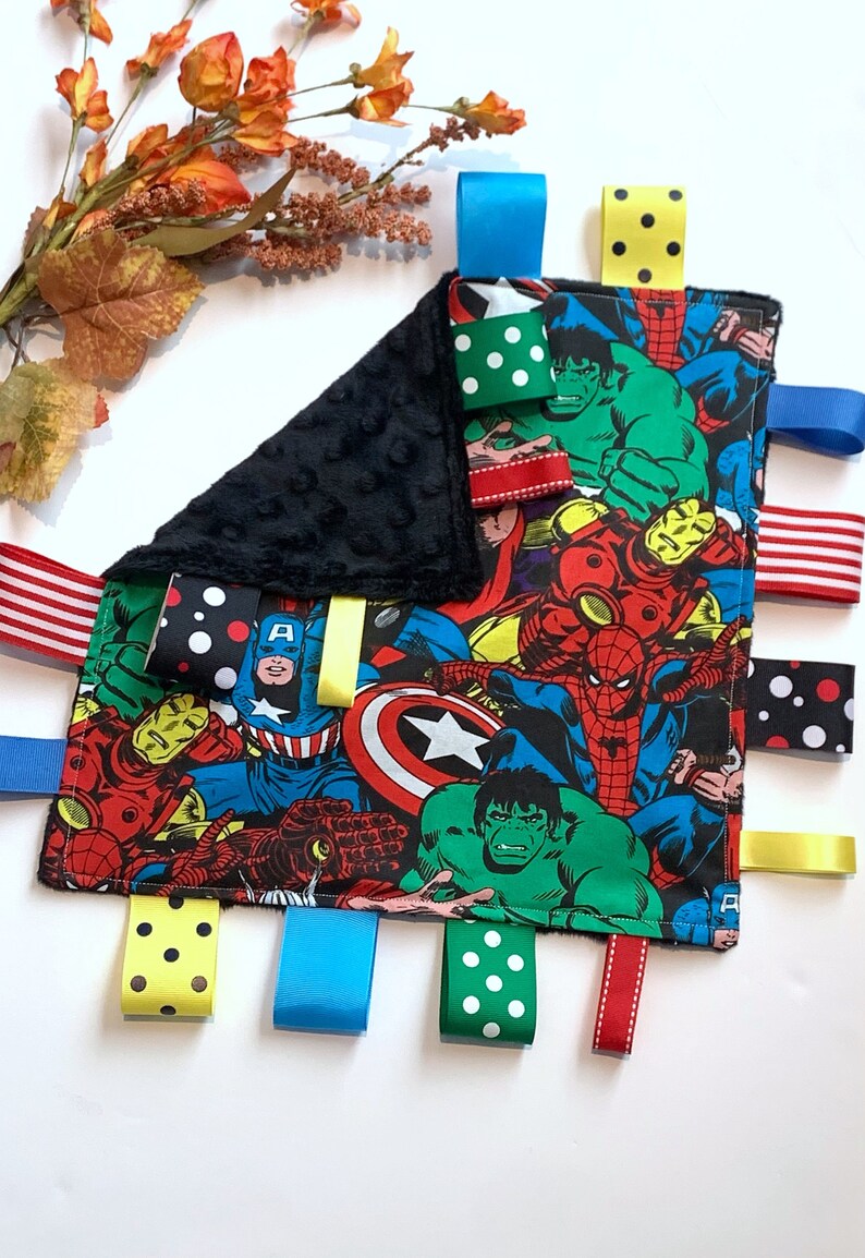 Op de afbeelding: Een zwart-wit babydeken met een kleurrijke superheldenprint. De deken heeft meerdere stoffen labels in verschillende kleuren en patronen, waaronder stippen, strepen en effen kleuren.