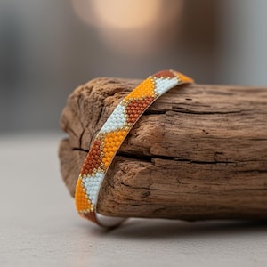 Puede incluir: Una pulsera de cuentas con un patrón geométrico en tonos naranja, marrón, blanco y dorado. La pulsera descansa sobre un trozo de madera flotante, mostrando el intrincado trabajo de cuentas y la paleta de colores. La pulsera mide aproximadamente 1,3 cm de ancho.
