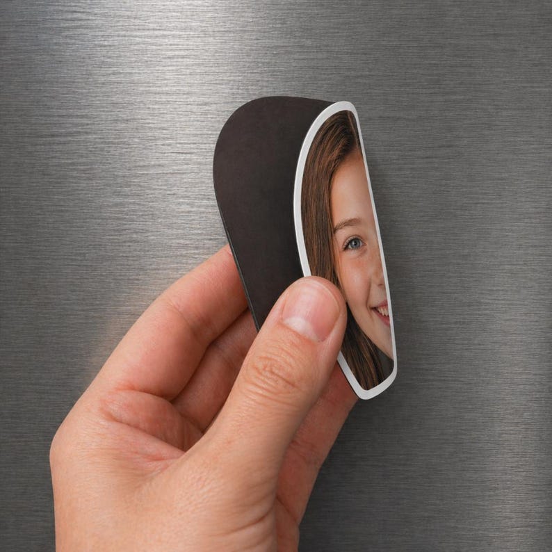 May include: A custom photo magnet featuring a portrait of a person with long brown hair. The magnet has a curved, semi-circular shape with a black back and a white border surrounding the photograph. It is shown attached to a brushed metal surface.