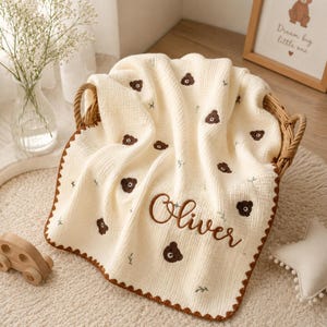 Può includere: Una coperta in mussola color crema con un motivo ripetuto di piccole facce di orso marroni e rametti verdi. La coperta è personalizzata con il nome Oliver ricamato in corsivo marrone e rifinita con un bordo smerlato di colore marrone.