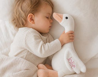 May include: A plush white goose toy with a soft texture, featuring a light peach-colored beak and feet. The name "jolie" is embroidered in pink thread on the wing of the goose. The toy has a simple, rounded body and a small black eye.