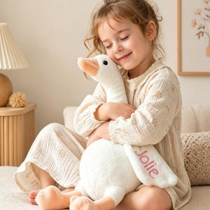 May include: A white plush goose toy with a long neck and orange feet, featuring the name Jolie embroidered in pink cursive script on one of its wings. The stuffed animal has a soft, fuzzy texture and black bead eyes, designed as a huggable toy.