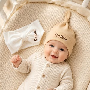 Könnte beinhalten: Eine beige Baby-Knotenmütze und ein weißes Stoff-Stirnband, beide mit dem Namen Kolbie in braunem Garn bestickt. Die Mütze ist in einem weichen, einfarbigen Beige mit einem Knoten oben gehalten, während das weiße Stirnband eine große, strukturierte Schleife aufweist.