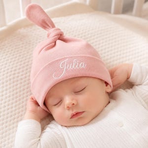 Könnte beinhalten: Eine zartrosa Babymütze mit einem Knoten an der Oberseite und einem umgeschlagenen Rand. Der Name Julia ist in weißer Schreibschrift auf der Vorderseite des Randes eingestickt. Die Mütze besteht aus glattem Stoff und sitzt eng am Kopf.