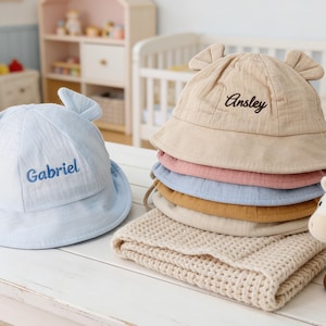 Può includere: Una pila di cappelli da sole per bambini in vari colori, tra cui blu, rosa e beige, con dettagli a forma di orecchie. Un cappello blu ha il nome "Gabriel" ricamato, e un cappello beige mostra "Ansley". Un unicorno di peluche e una coperta color crema sono anche presenti.