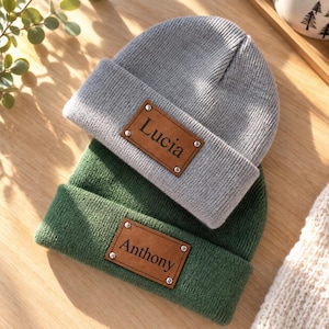 May include: A light gray knit beanie and a dark green knit beanie, each featuring a rectangular brown leather patch secured with four silver-toned metal rivets. The gray beanie has the name Lucia embossed, and the green beanie has Anthony.