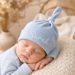 May include: A light blue baby hat with a top knot and the name Sarah embroidered in white cursive script on the front. The hat is made of a soft fabric and is worn by a sleeping infant dressed in a matching light blue long-sleeve waffle knit shirt.