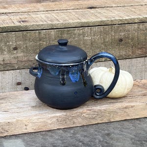 Może przedstawiać: Matowy czarny ceramiczny garnek z pokrywką, zaokrąglonym korpusem, dwoma małymi uchwytami bocznymi i dużym zakrzywionym uchem. Górna krawędź posiada dekoracyjny wzór niebiesko-białych zacieków szkliwa. Za garnkiem znajduje się mała biała dynia.