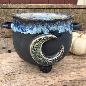 May include: A black ceramic cauldron with two side handles and three small feet. The rim features a blue and white drip glaze, and the front is decorated with a raised, textured crescent moon design with intricate swirling patterns.