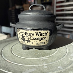 May include: A matte black ceramic cauldron with a lid, two side handles, and three small feet. A cream-colored label on the front features a silhouette of a witch on a broomstick and the text: Pure Witch Essence. USE: LATHER ON TO GROW HAIR.