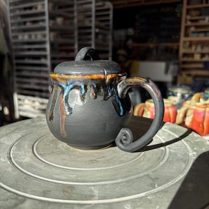 May include: A matte black ceramic mug with a matching lid, featuring a decorative drip glaze in shades of metallic blue, copper, and bronze around the rim. The mug has a curved handle that ends in a distinct spiral scroll at the base.