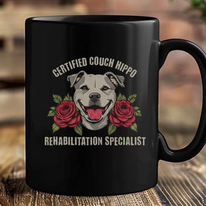 Op de afbeelding: Een zwarte keramische mok met een afbeelding van een grijs pitbull-hondengezicht in het midden tussen twee rode rozen. De tekst boven de hond luidt CERTIFIED COUCH HIPPO en de tekst onder de hond luidt REHABILITATION SPECIALIST in lichtbeige.