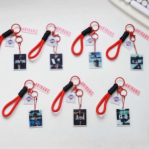 May include: A collection of keychains with red wrist straps and black accents. Each keychain features a small, clear square with a photo of a person and the name of the person. The keychains also have a tag that says "ARIRANG".