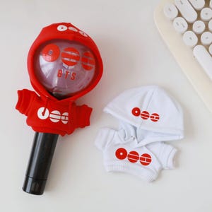May include: A red and white light stick cover and a matching miniature hoodie. The red hoodie has white circular and horizontal line designs and the text "BTS". The black light stick has the text "THE SOUL".