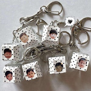 Pode incluir: Uma coleção de chaveiros personalizados. Cada chaveiro apresenta o rosto de uma criança em um cubo branco adornado com padrões de estrelas pretas. Possuem fechos e correntes prateadas.