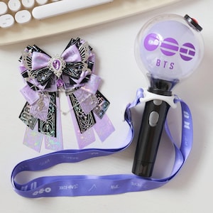 May include: A purple and black bow with a jeweled centre and dangling charms, next to a black and white BTS light stick with a purple strap. The light stick has the letters "BTS" printed on it.